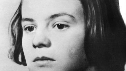 Undatierte Porträtaufnahme von Sophie Scholl. Sie hat dunkle Haare und trägt einen Seitenscheitel, ihr Oberteil ist auch von dunkler Farbe. Undatierte Porträtaufnahme von Sophie Scholl. Sie hat dunkle Haare und trägt einen Seitenscheitel, ihr Oberteil ist auch von dunkler Farbe.