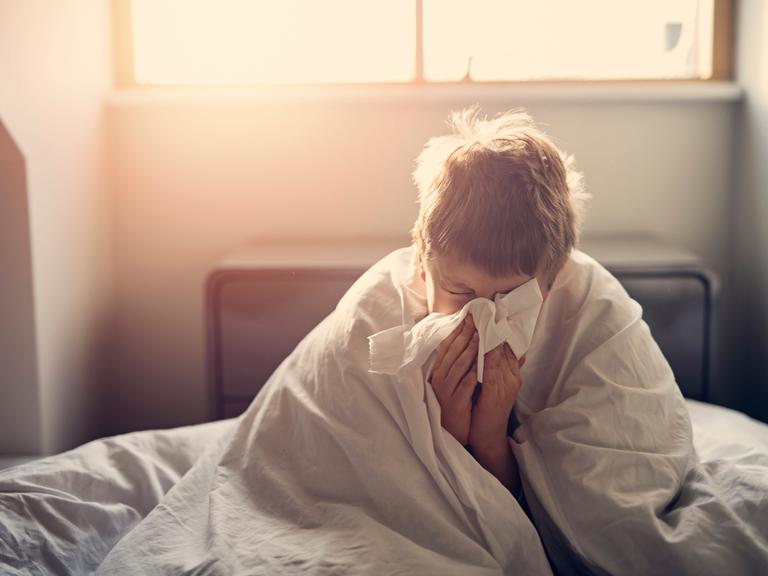 Ein kleiner Junge liegt krank im Bett mit einem Taschentuch vor seiner Nase.