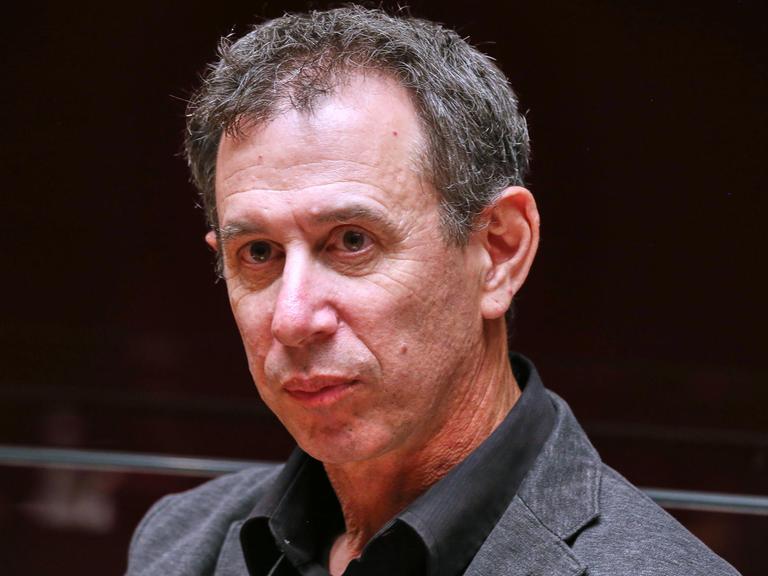 Portrait von Assaf Gavron, der vor einem dunklen Hintergrund sitzt und an der Kamera vorbeischaut.