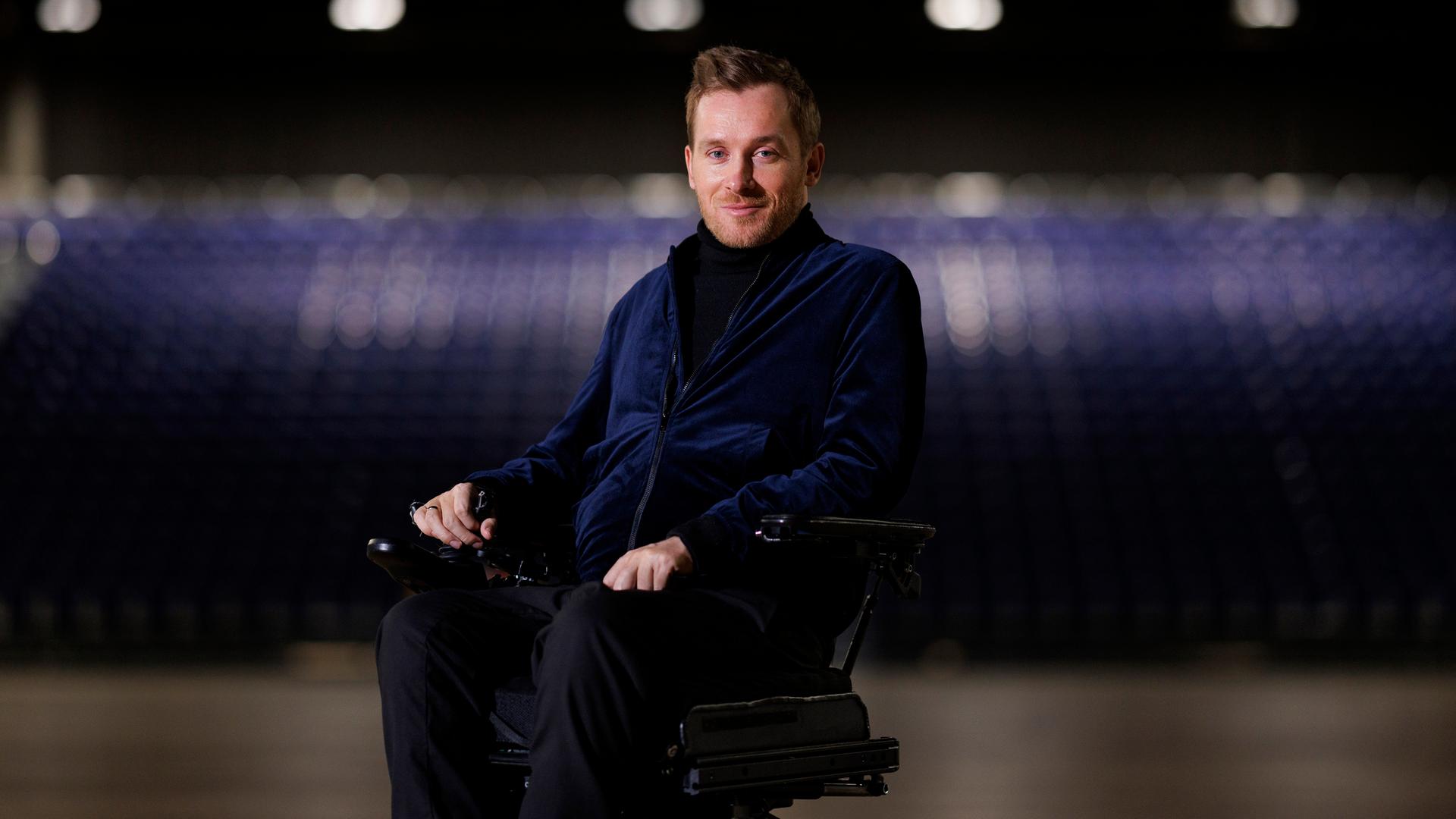 Der querschnittsgelähmte Schauspieler Samuel Koch sitzt in einem Rollstuhl in einer großen Halle. Im Hintergrund sieht man verschwommen mehrere Sitzreihen. 