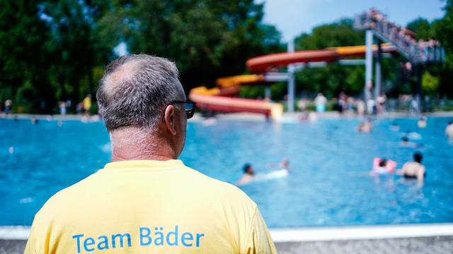 Ein Bade-Meister in einem gelben Hemd steht am Backen-Rand. Im Schwimm-Becken sind mehrere Menschen.