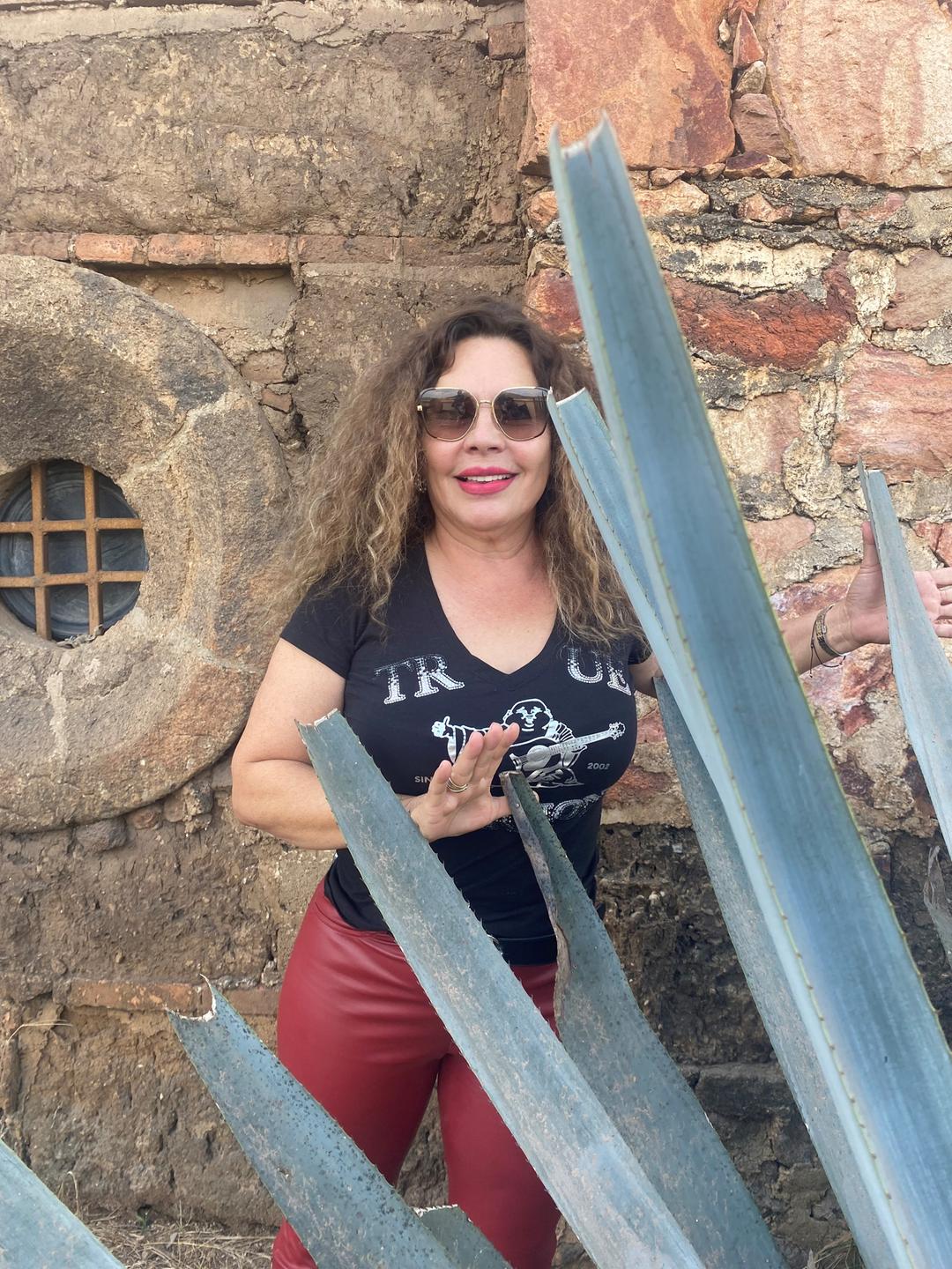Die Brennereibesitzerin Leticia Hermosillo, mit langen lockigen Haaren, Sonnenbrille und roter Lederhose, steht hinter einer großen Agavenpflanze vor einer Mauer.