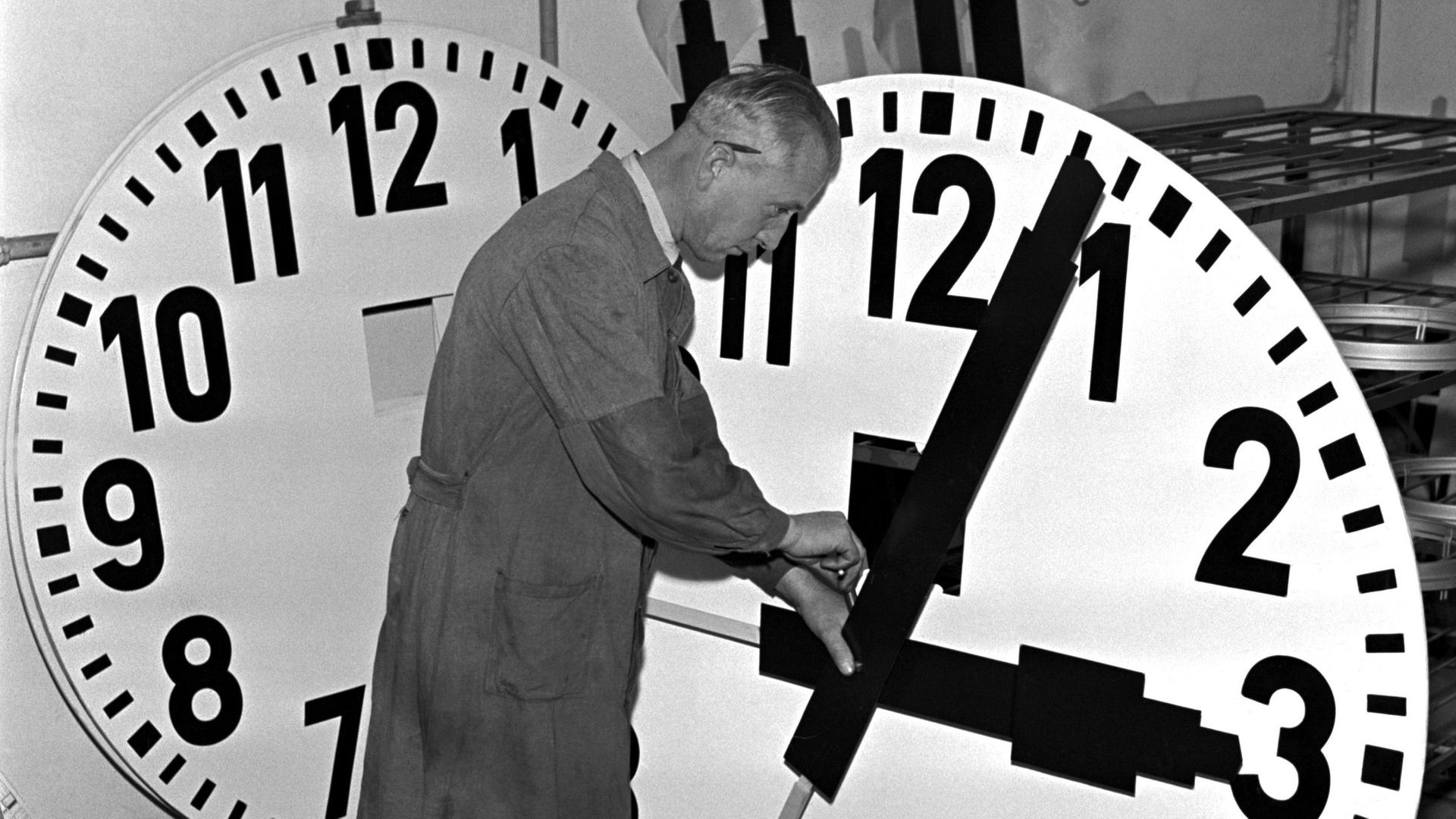 Historisches Schwarzeißfoto eines Uhrmachers im Arbeitskittel, der mit einem großen Ziffernblatt hantiert.