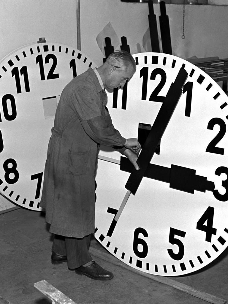 Historisches Schwarzeißfoto eines Uhrmachers im Arbeitskittel, der mit einem großen Ziffernblatt hantiert.