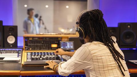Ein Tontechniker mit Kopfhörern bedient ein Mischpult in einem Aufnahmestudio, im Hintergrund sieht man unscharf einen Sänger in einem Aufnahmeraum, der in ein Mikrofon singt.
