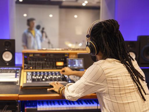 Ein Tontechniker mit Kopfhörern bedient ein Mischpult in einem Aufnahmestudio, im Hintergrund sieht man unscharf einen Sänger in einem Aufnahmeraum, der in ein Mikrofon singt.