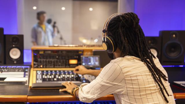 Ein Tontechniker mit Kopfhörern bedient ein Mischpult in einem Aufnahmestudio, im Hintergrund sieht man unscharf einen Sänger in einem Aufnahmeraum, der in ein Mikrofon singt.
