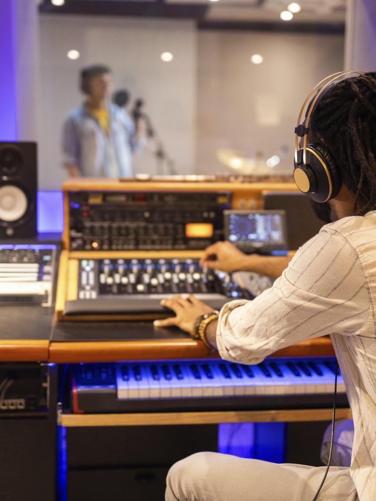 Ein Tontechniker mit Kopfhörern bedient ein Mischpult in einem Aufnahmestudio, im Hintergrund sieht man unscharf einen Sänger in einem Aufnahmeraum, der in ein Mikrofon singt.