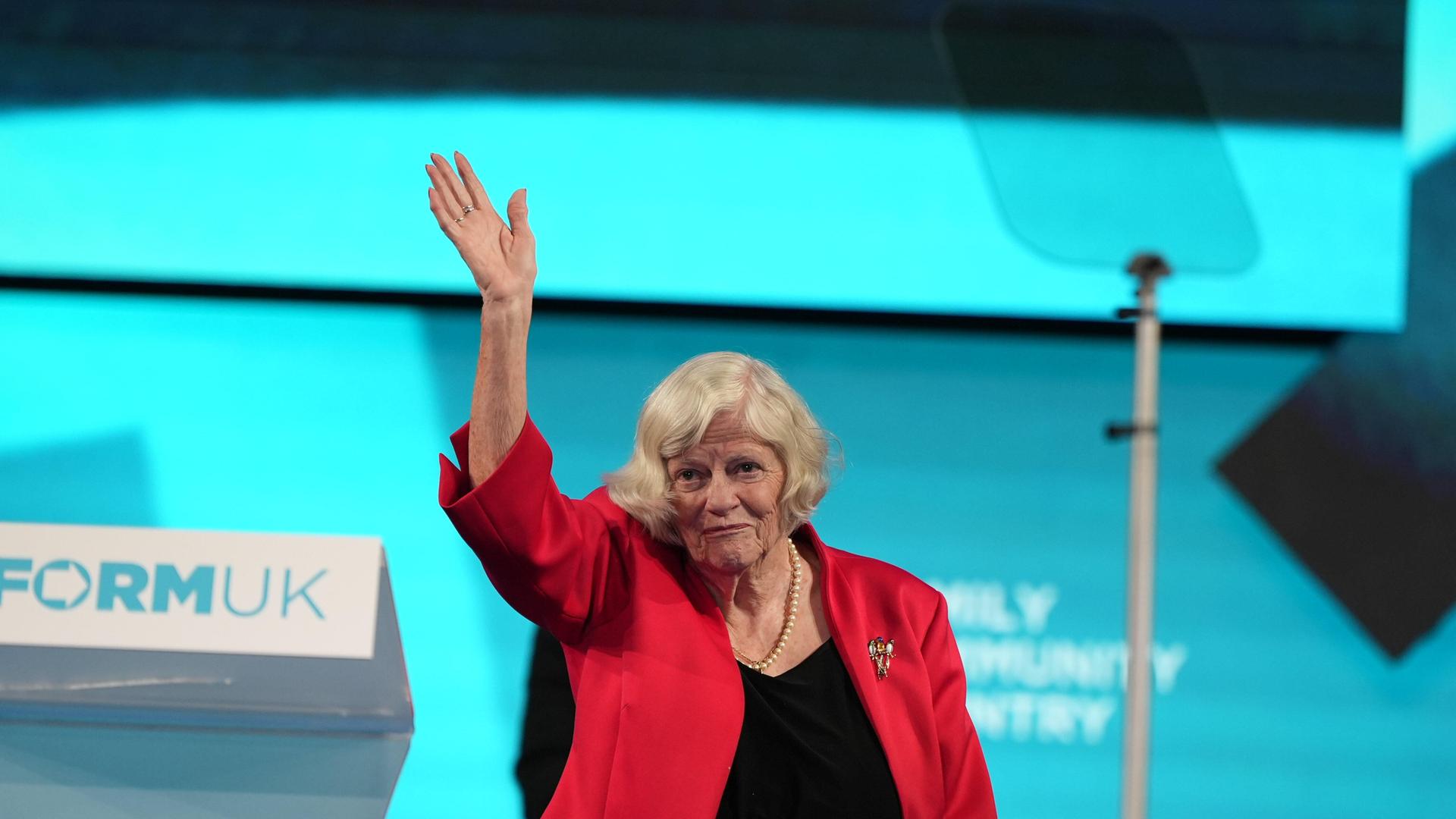 Das Bild zeigt die Politikerin Ann Widdecombe bei einer Rede auf einer Bühne. Sie trägt einen roten Blazer und winkt ins Publikum. Im Hintergrund ist ein Rednerpult mit der Aufschrift "Reform UK" zu erkennen.