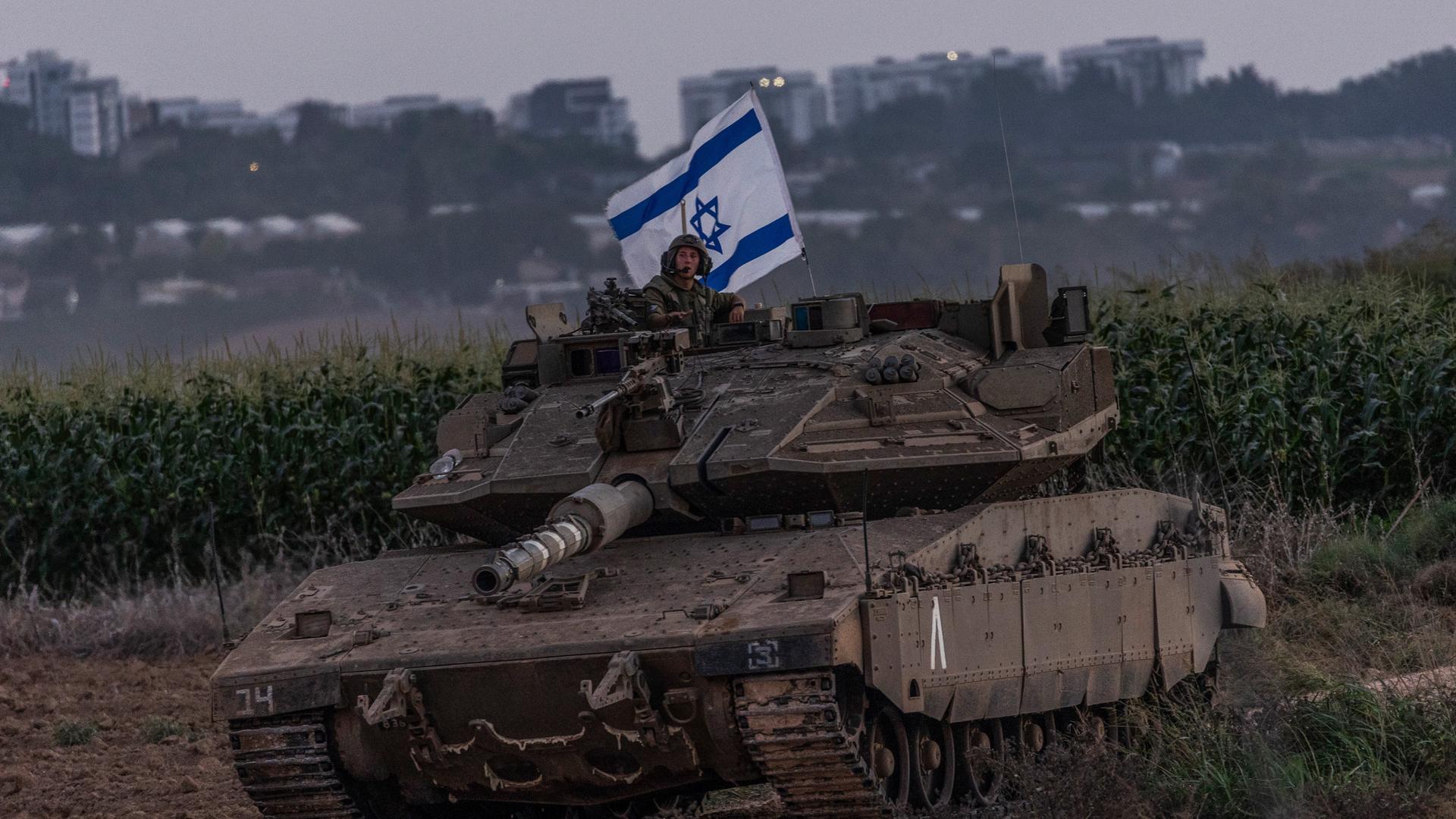 Ein Panzer, daraus schaut ein Soldat mit israelischer Flagge. Im Hintergrund ist besiedeltes Gebiet zu erkennen. 