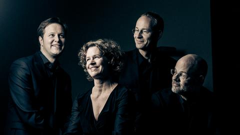 Die vier Musiker, eine Frau umringt von drei Männern, lächlel versonnen, während sie eng in dunkler Kleidung beieinander sitzen.