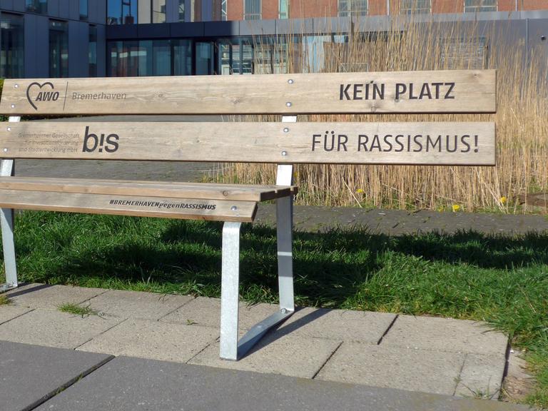 Die Rückenlehne einer Holzbank ragt über die Sitzfläche hinaus. Dort, wo niemand sitzen kann, steht auf der Lehne: "Kein Platz für Rassismus".