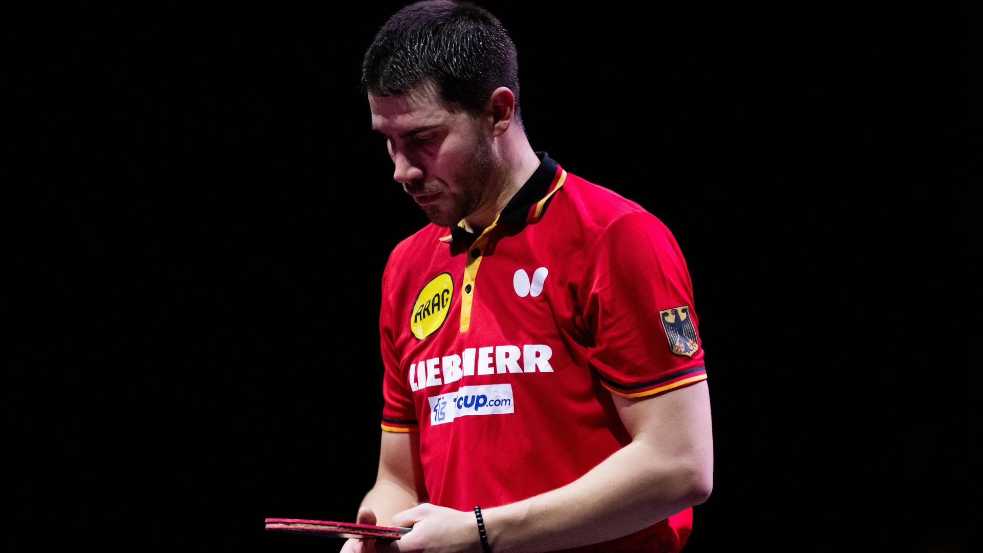 Patrick Franziska schaut nach unten und hält einen Tischtennisschläger in den Händen. Er trägt ein rotes Trikot. Patrick Franziska schaut nach unten und hält einen Tischtennisschläger in den Händen. Er trägt ein rotes Trikot.