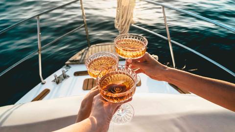 Auf dem Deck eines fahrenden Schiffes wird mit drei Champagnerschalen angestoßen. Auf dem Deck eines fahrenden Schiffes wird mit drei Champagnerschalen angestoßen.