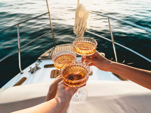 Auf dem Deck eines fahrenden Schiffes wird mit drei Champagnerschalen angestoßen. Auf dem Deck eines fahrenden Schiffes wird mit drei Champagnerschalen angestoßen.