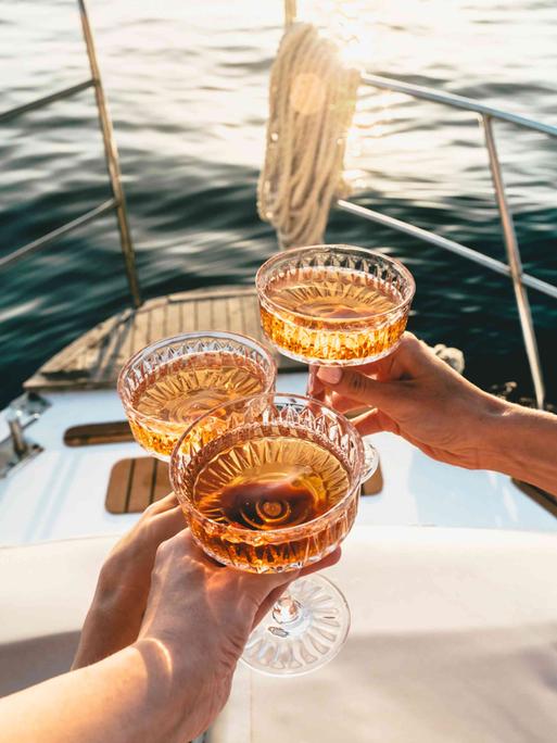 Auf dem Deck eines fahrenden Schiffes wird mit drei Champagnerschalen angestoßen. Auf dem Deck eines fahrenden Schiffes wird mit drei Champagnerschalen angestoßen.