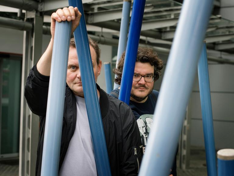 Devid Striesow und Axel Ranisch stehen unter einem Klettergerüst und schauen ernst in die Kamera