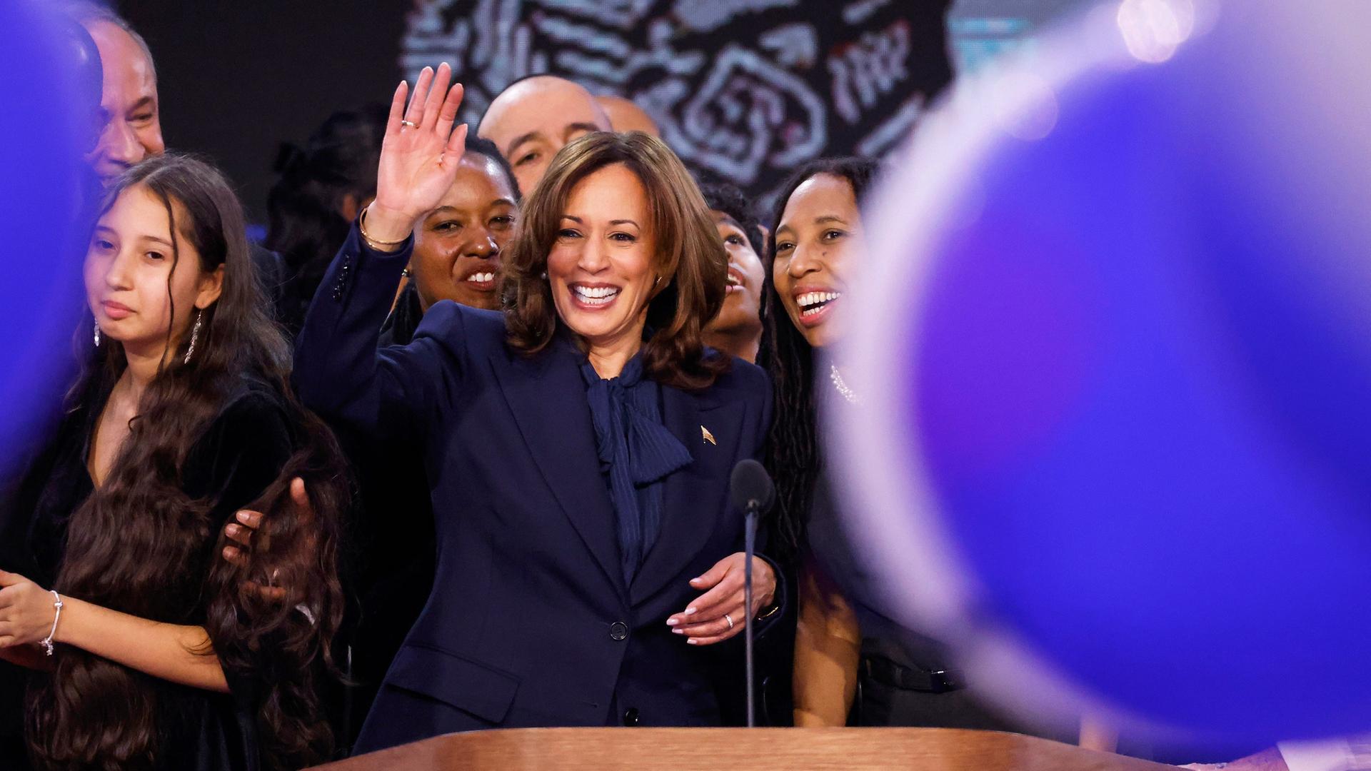 Kamala Harris steht hinter einem Rednerpult und winkt. Neben und hinter ihr stehen Familie und Parteifreunde. Im Vordergrund sieht man blaue Luftballons.