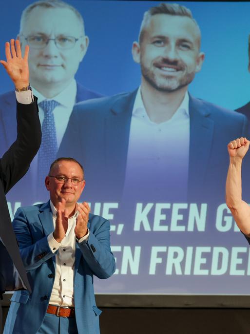AfD-Politiker Ulrich Siegmund (links), Tino Chrupalla und Kabarettist Uwe Steimle stehen lächelnd und klatschend auf einer Bühne, dahinter ist ein großes Foto mit den dreien zu sehen AfD-Politiker Ulrich Siegmund (links), Tino Chrupalla und Kabarettist Uwe Steimle stehen lächelnd und klatschend auf einer Bühne, dahinter ist ein großes Foto mit den dreien zu sehen