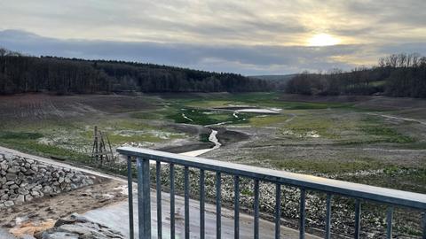 Blick vom Staudamm aus in die leere Steinbachtalsperre. Darin sieht man einen kleinen Wasserlauf und Pfützen. Im Vordergrund steht ein Geländer.