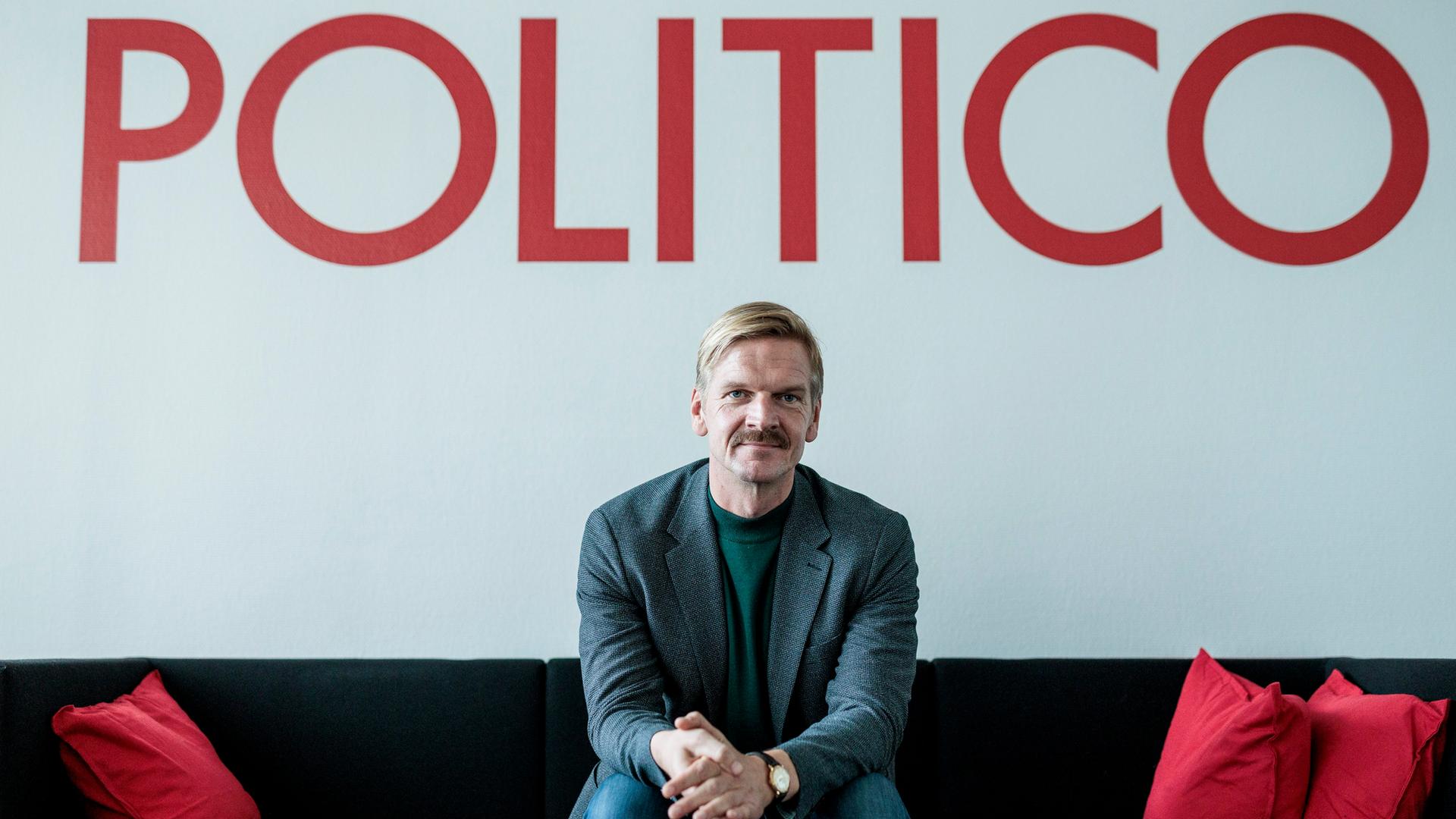 Gordon Repinski, Chefredakteur des Nachrichtenportals "Politico" sitzt auf einem Sofa unter einem roten Schriftzug der Medienmarke.