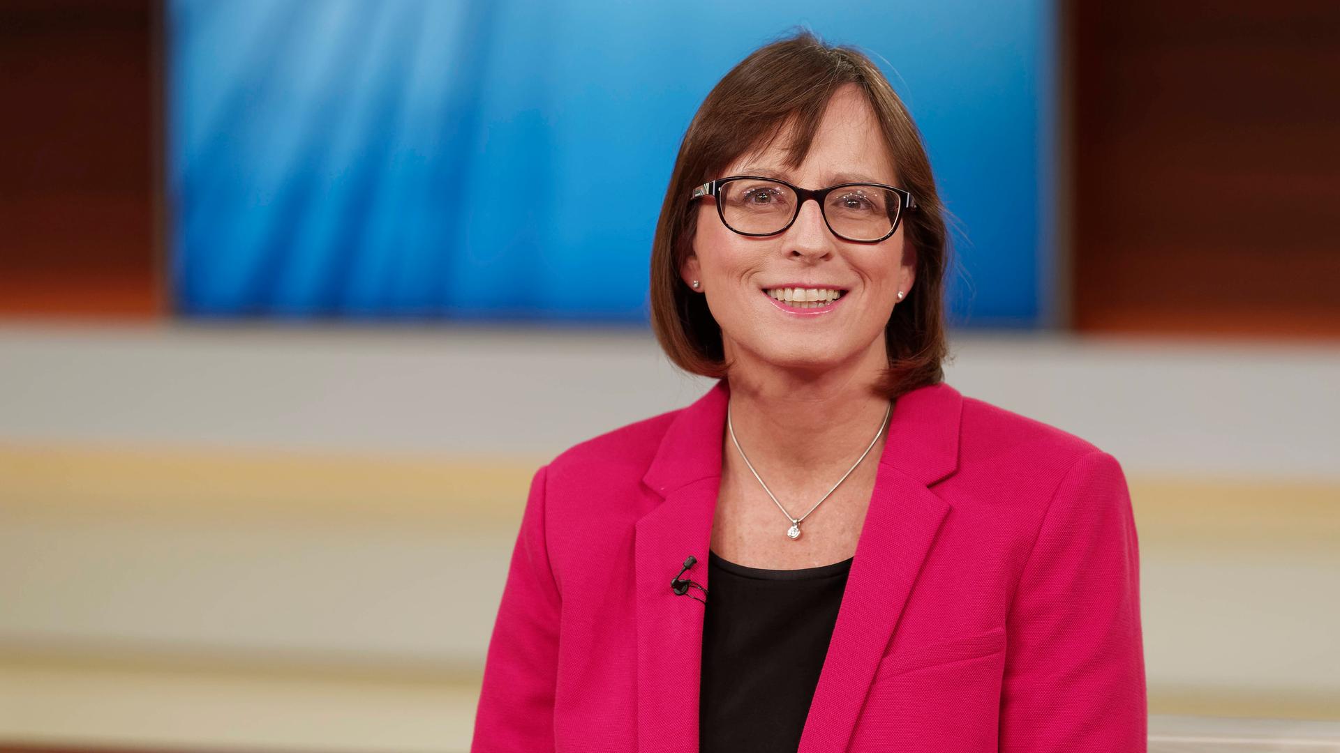 Britta Waldschmidt-Nelson sitzt in einem Fernsehstudio und schaut freundlich in die Kamera. Sie trägt Brille und ein pinkfarbenes Sakko.