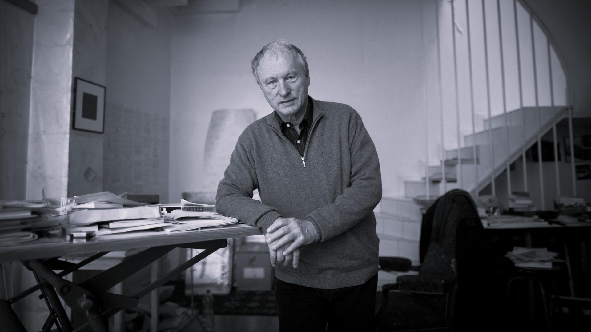 Schwarzweiß-Aufnahme von Valère Novarina. Er steht an einem Schreibtisch und stützt seinen Ellenbogen auf die Tischplatte.
