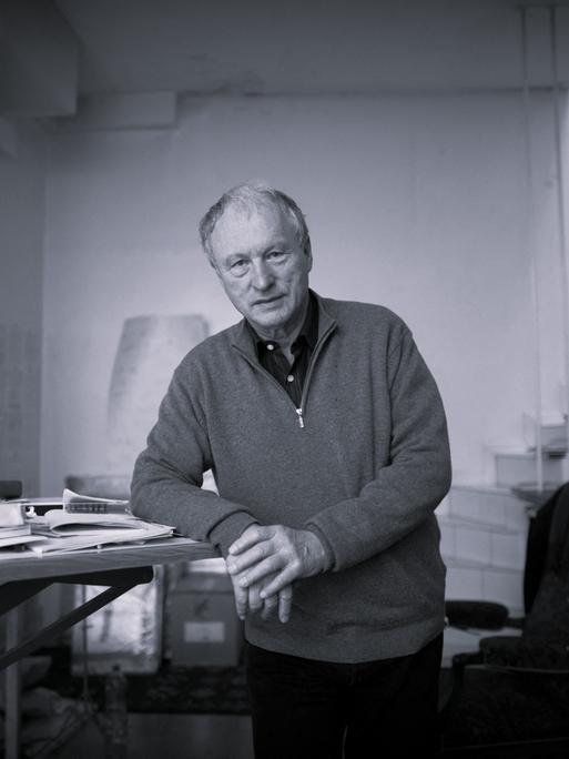 Schwarzweiß-Aufnahme von Valère Novarina. Er steht an einem Schreibtisch und stützt seinen Ellenbogen auf die Tischplatte.