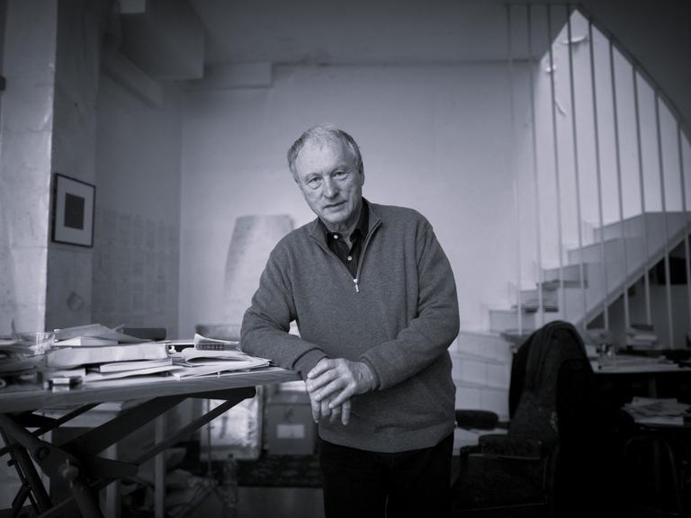 Schwarzweiß-Aufnahme von Valère Novarina. Er steht an einem Schreibtisch und stützt seinen Ellenbogen auf die Tischplatte.