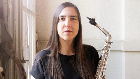 Eine Frau mit braunen, glatten Haaren blickt ernst in die Kamera. Sie trägt ein schwarzes T-Shirt und hält ein Saxophon. Im Hintergrund ist ein Fenster zu sehen.