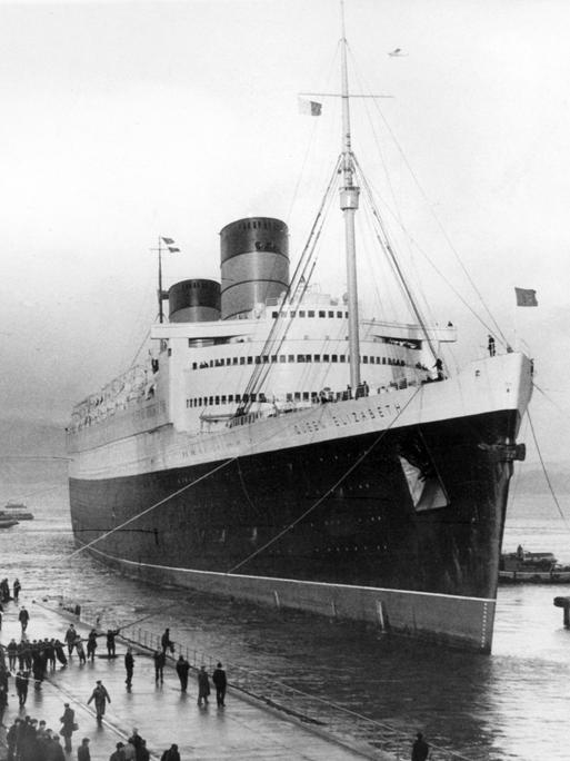 Schwarz-Weiß-Aufnahme des damals größten Passagierschiff der Welt, der Queen Elizabeth der Cunard Line, in ein Trockendock im Firth of Clyde in Schottland. Mehrere Arbeiter leinen das Schiff an.