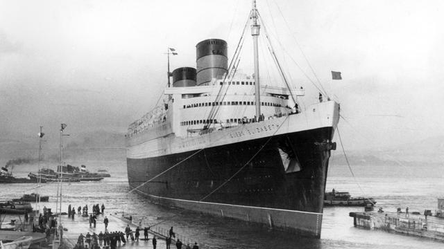 Schwarz-Weiß-Aufnahme des damals größten Passagierschiff der Welt, der Queen Elizabeth der Cunard Line, in ein Trockendock im Firth of Clyde in Schottland. Mehrere Arbeiter leinen das Schiff an.