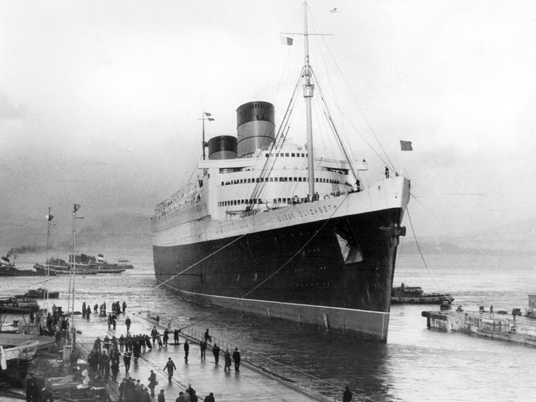 Schwarz-Weiß-Aufnahme des damals größten Passagierschiff der Welt, der Queen Elizabeth der Cunard Line, in ein Trockendock im Firth of Clyde in Schottland. Mehrere Arbeiter leinen das Schiff an.