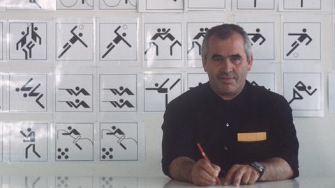 Der Grafiker, Designer, Architekt, Otl Aicher (1922-1991) sitzt 1970 an seinem Schreibtisch, im Hintergrund eine Tafel mit Piktogrammen, Symbolen, Zeichen, die er für die Sportarten der Olympischen Sommerspiele 1972 in München entworfen hat.