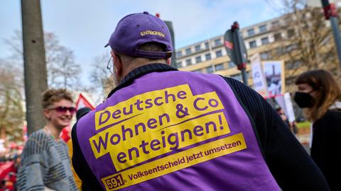 Auf einer Demonstration steht ein Mann, der eine West der Initiative "Deutsche Wohnen & Co. enteignen" trägt. Er ist von hinten fotografiert. Die Weste ist in den Farben lila und gelb gehalten. Auf ihr steht unter "Deutsche Wohnen & Co. enteignen" der Slogan "59,1 % - Volksentscheid jetzt umsetzen"