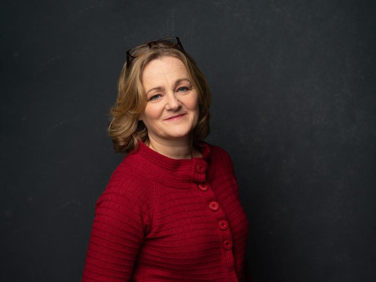 Die deutsch-norwegische Kinderpsychologin Katrin Glatz Brubakk steht mit roter Strickjacke vor dunklem Hintergrund.