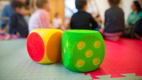 Zwei Würfel liegen in einer Kindertagesstätte vor Kindern, die in einem Kreis sitzen.