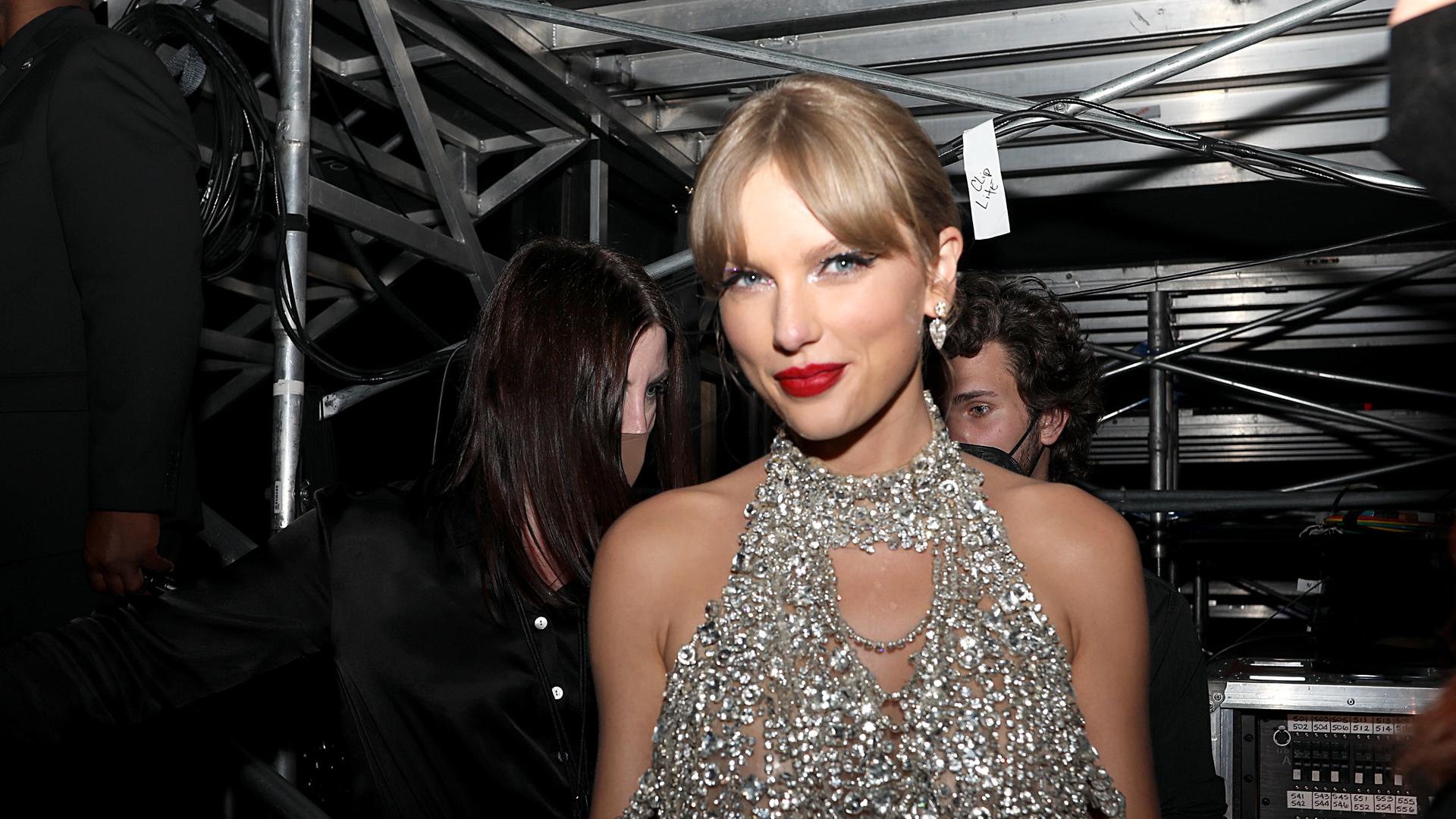 Taylor Swift hinter der Bühne in silber glänzendem Kleid vor schwarzem Hintergrund.