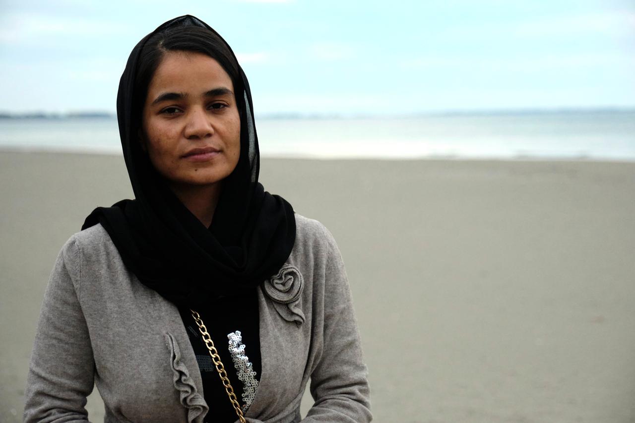 Ziagul Azimi steht am Strand vor dem Wasser. Sie trägt ein schwarzes Kopftuch und eine graue Jacke.