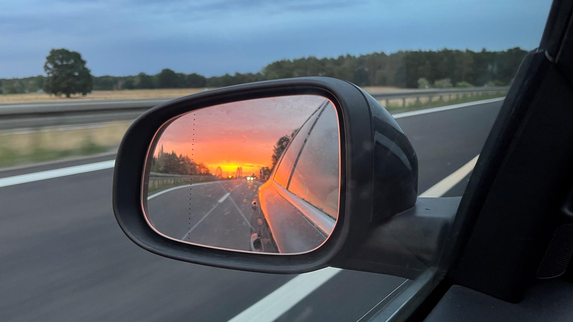 Aufnahme eine Rückspiegels in dem sich die aufgehende Sonne spiegelt. Das Auto fährt auf einer Autobahn.