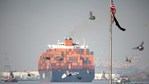 Ein Container-Schiff fährt in den Hafen von New Jersey in den USA ein, rechts im Vordergrund ist ein Flaggenmast mit herunterhängender US-Fahne.
