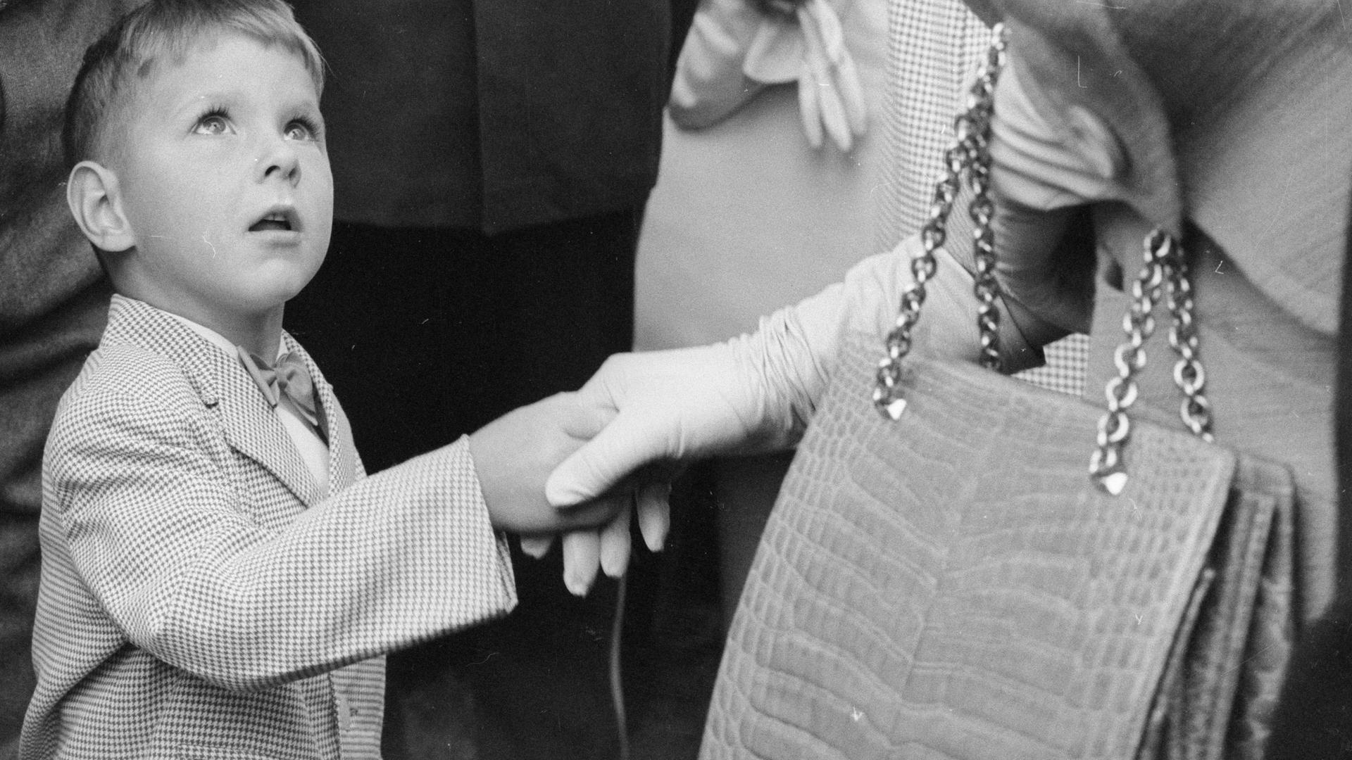 Historisches Schwarzweißfoto eines kleinen Jungen, der die Hand einer deutlich größeren und elegant gekleideten Frau schüttelt und staunend zu ihr aufschaut.