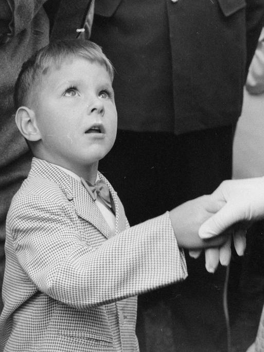 Historisches Schwarzweißfoto eines kleinen Jungen, der die Hand einer deutlich größeren und elegant gekleideten Frau schüttelt und staunend zu ihr aufschaut. Historisches Schwarzweißfoto eines kleinen Jungen, der die Hand einer deutlich größeren und elegant gekleideten Frau schüttelt und staunend zu ihr aufschaut.