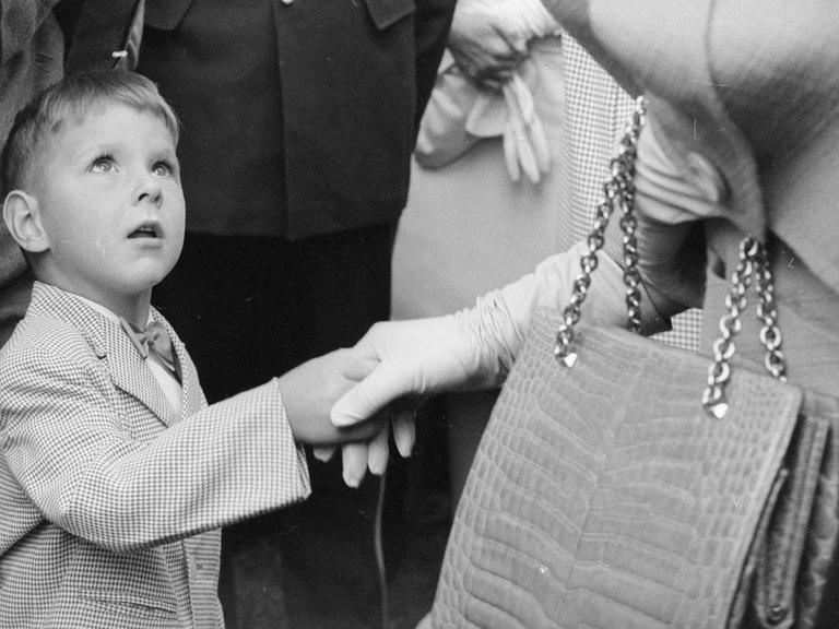 Historisches Schwarzweißfoto eines kleinen Jungen, der die Hand einer deutlich größeren und elegant gekleideten Frau schüttelt und staunend zu ihr aufschaut. Historisches Schwarzweißfoto eines kleinen Jungen, der die Hand einer deutlich größeren und elegant gekleideten Frau schüttelt und staunend zu ihr aufschaut.