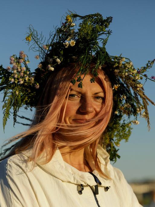 Eine Frau in weißer Tracht lächelt in die Kamera. Sie feiert die Mittsommernacht und trägt Zweige und Blätter im Haar. 