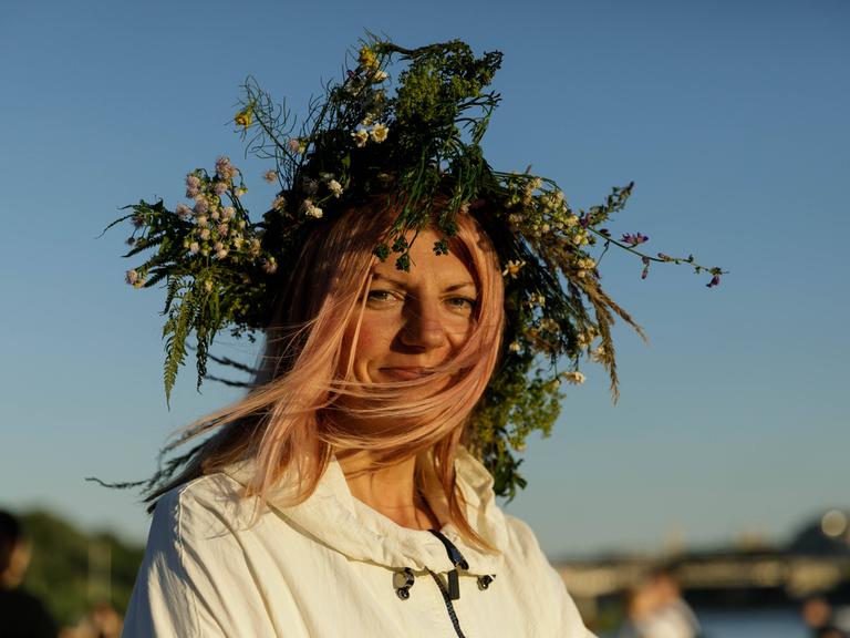 Eine Frau in weißer Tracht lächelt in die Kamera. Sie feiert die Mittsommernacht und trägt Zweige und Blätter im Haar. 