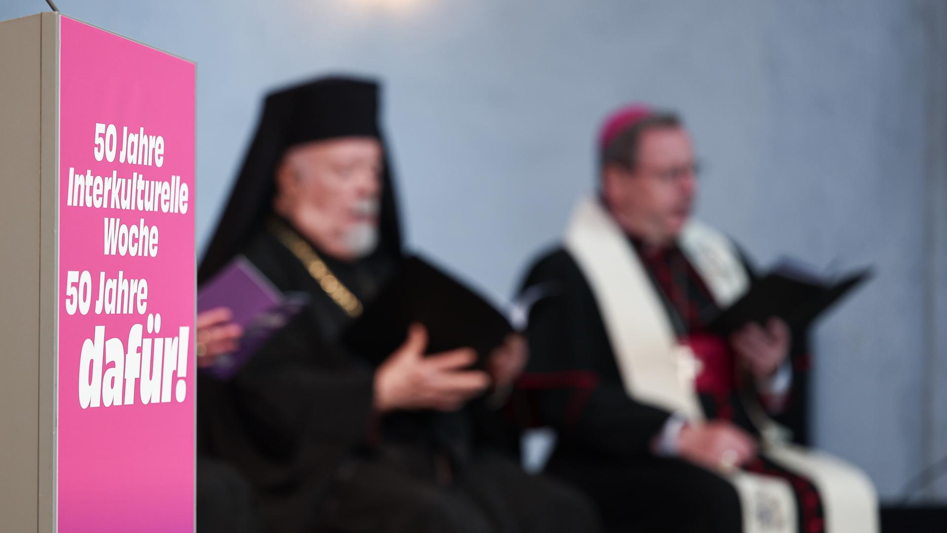 Im Vordergrund ein magantafarbenes Schild mit der Aufschrift "50 Jahre Interkulturelle Woche". Dahinter unscharf je ein sitzender Geistlicher der griechisch-orthodoxen und der katholischen Kirche. 