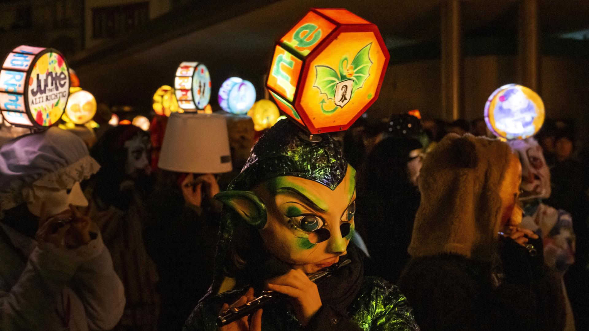 Menschen mit großen bunten Masken ziehen mit einer bunten Laternen umher.