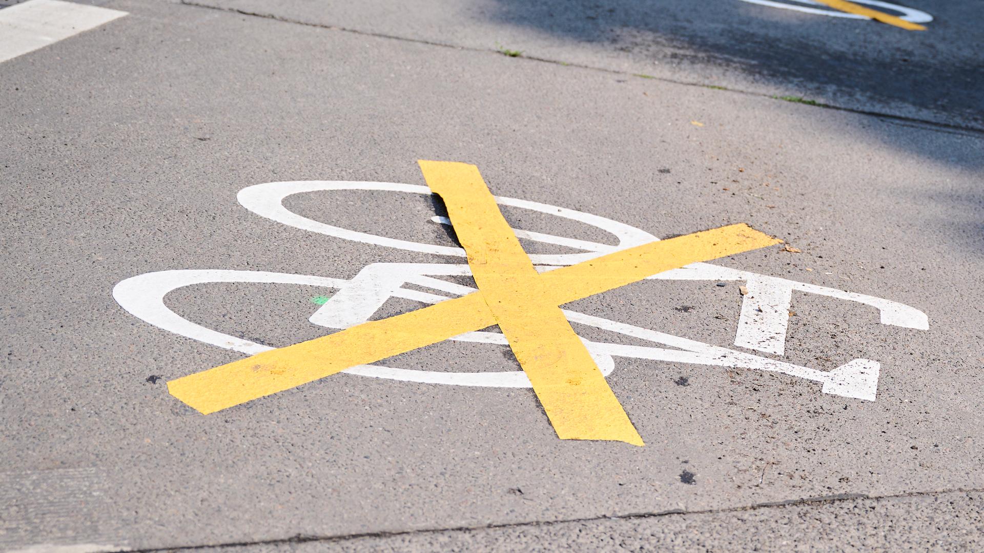 Auf einer Straße sind die Zeichen für den Radweg mit gelben Kreuzen zugeklebt, und auf dem früheren Radweg parken Autos.