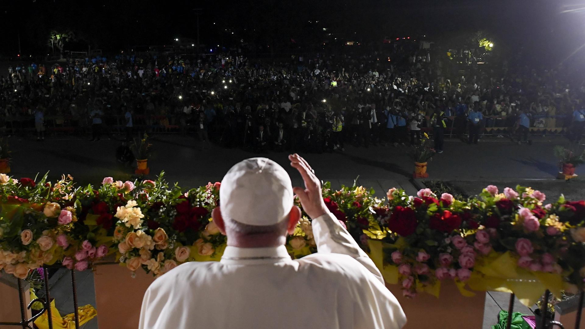 Papst Franziskus steht in Papua-Neuguinea auf einer beleuchteten Bühne vor einer Menschenmenge im Dunkeln, die Lichter in die Höhe hält. 